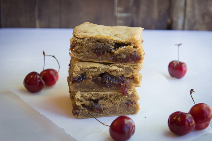 Cherry Jubilee Blondies stacked in a tower of three with a wood background and fresh cherries in the foreground